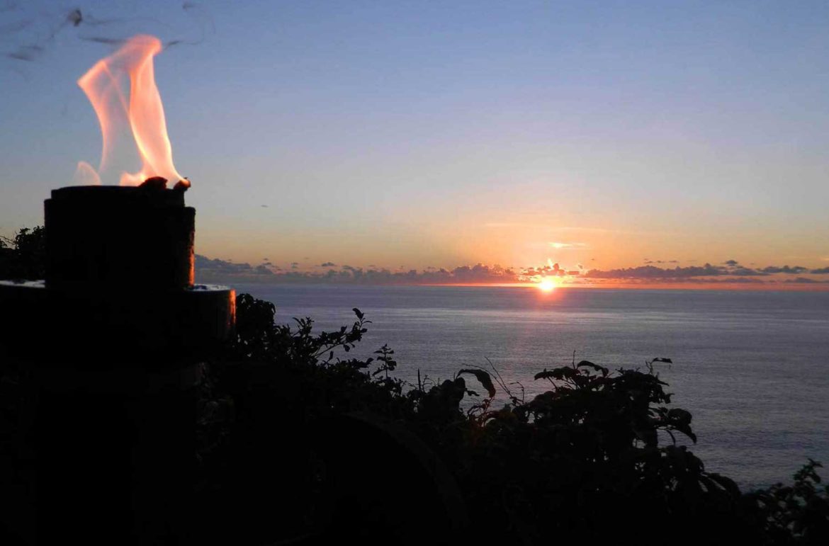 Chama acesa no Templo de Uluwatu, na Ilha de Bali (Indonésia), com o pôr do sol no mar ao fundo