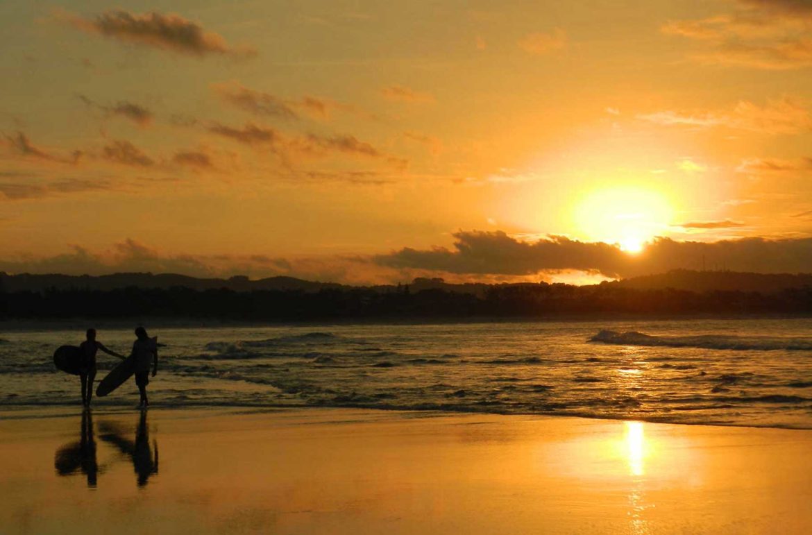 Casal de surfistas sai do mar ao pôr do sol na Praia The Pass, em Byron Bay (Austrália)