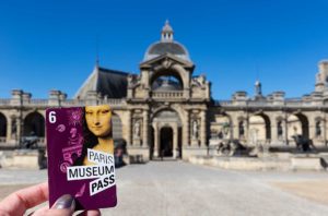 Mão segura o Paris Museum Pass em frente ao Castelo de Chantilly, na França