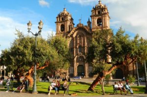 Pessoas sentam nos bancos da Plaza de Armas, em Cusco, com a Igreja da Compania de Jesus ao fundo