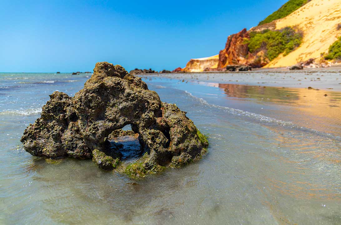 Pedra com furo na beira da Praia de Ponta Grossa, com as falésias ao fundo