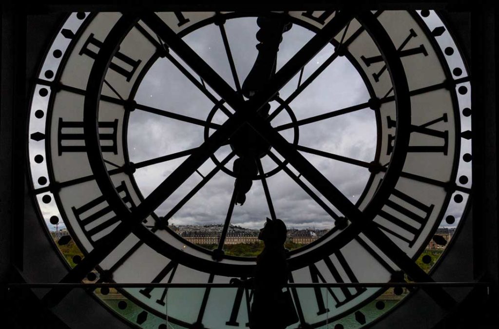 Visitante para para foto diante do relógio do quinto andar do Museu D'Orsay, com os céus de Paris ao fundo