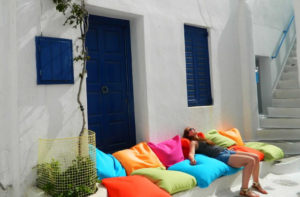 Mulher deita ao sol sobre almofadas coloridas no centro histórico de Mykonos