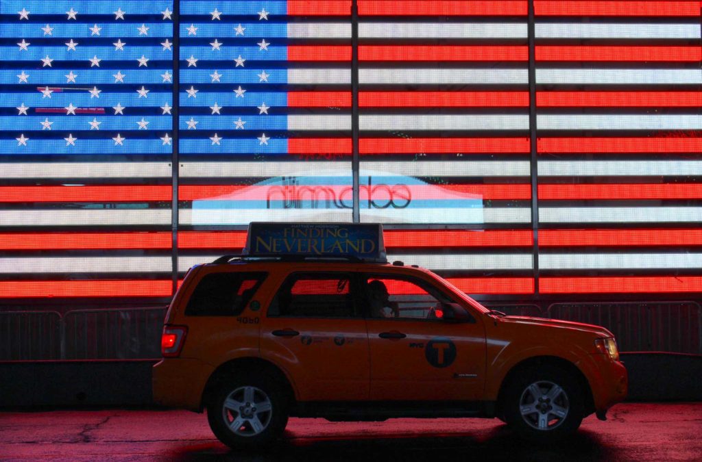 Táxi passa em frente a painel de luzes de neon que mostra a bandeira dos EUA na Times Square