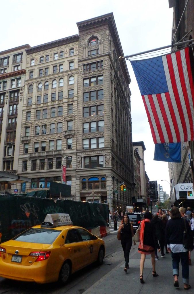 Pedestres caminham sob bandeira dos EUA em calçada de Nova York enquanto táxi passa pela rua