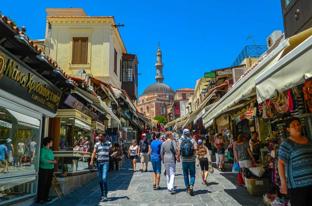 Pessoas caminham pelas ruelas de Rhodes com a Mesquita de Suleiman ao fundo