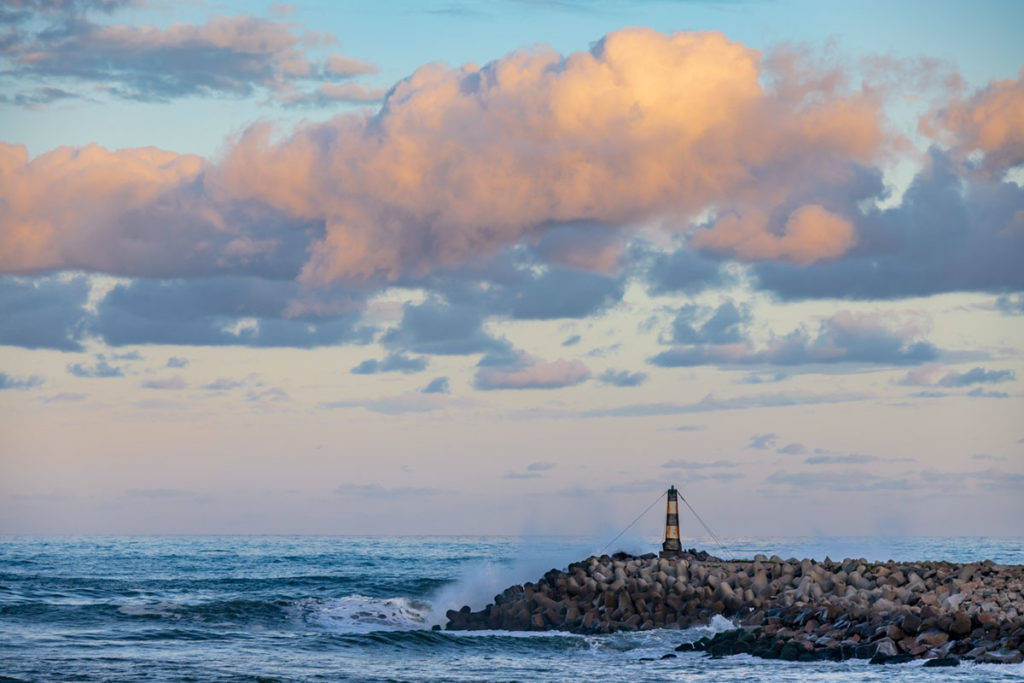 Ondas batem nos rochedos sob o farol dos Molhes da Barra ao entardecer