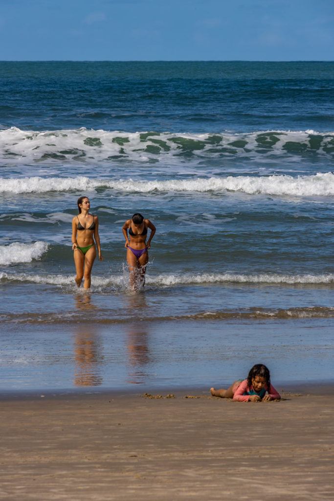 Mulheres saem do mar enquanto criança brinca na areia na Praia do Rosa